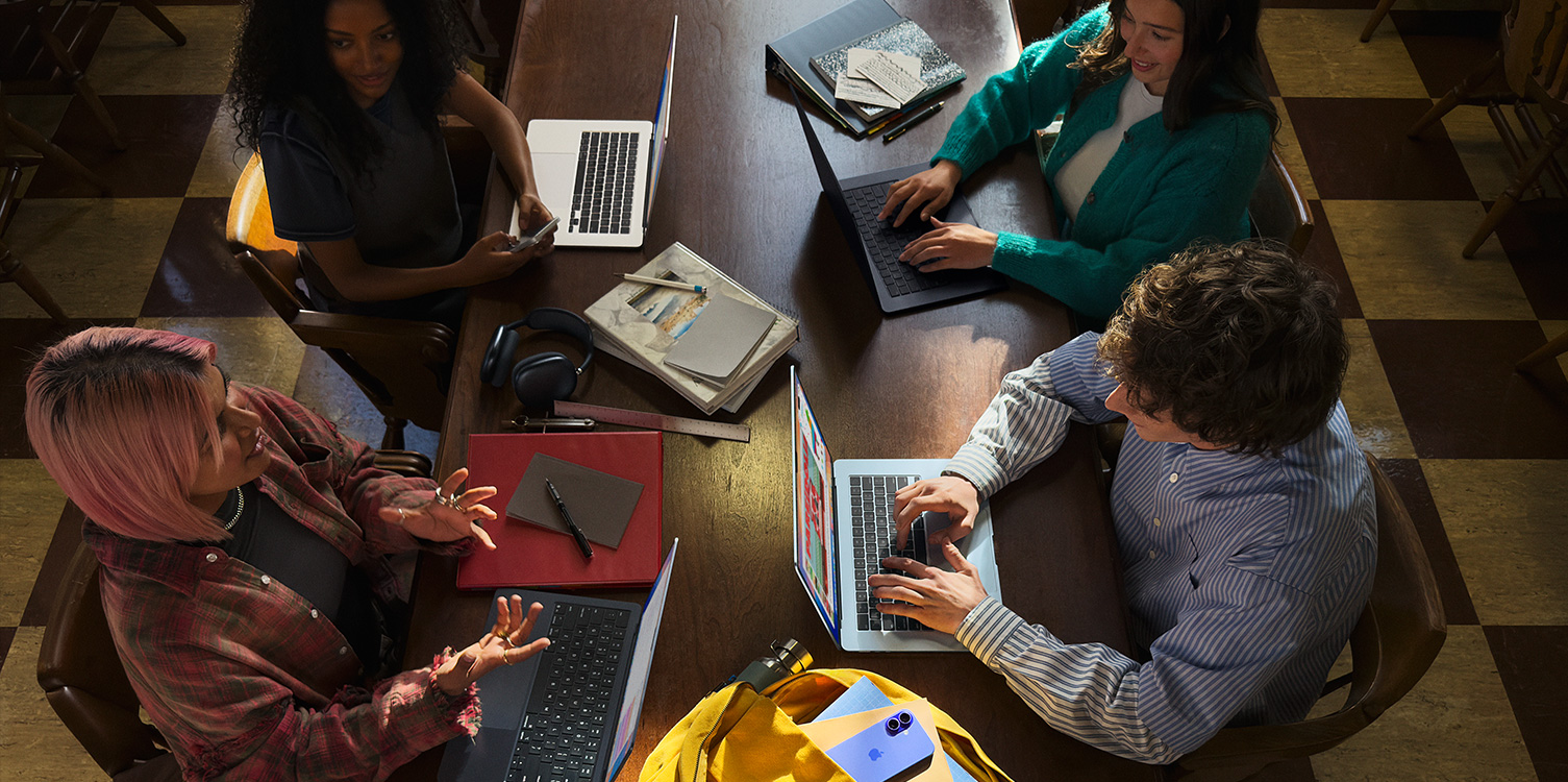 Empat mahasiswa duduk di dekat sebuah meja. Mereka sedang membuka MacBook dan catatan.