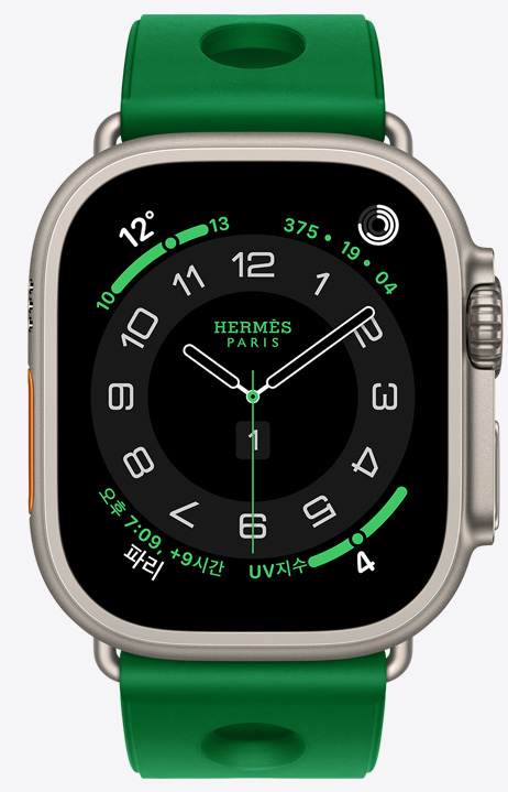 Apple Watch Hermès Ultra 4 Circulaire 아날로그 시계 페이스 및 시, 분, 초침, 컴플리케이션. 날씨, 활동 링, 파리 시간 및 자외선 지수가 표시된 모습. 오른쪽 옆면 외관에 Digital Crown과 돌출된 측면 버튼이 보이고, 베르 베흐티고(밝은 초록색) 색상의 성형 고무 소재에 중앙에 구멍이 뚫린 Scub’H Diving 밴드와 연결되어 있는 모습