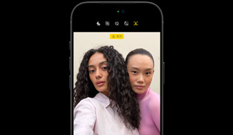 iPhone 18 Pro 블랙 모델이 앞을 향한 모습. 두 사람에 얼굴 추적 기능이 적용되고 있는 Center Stage 전면 카메라 기능을 보여줍니다.