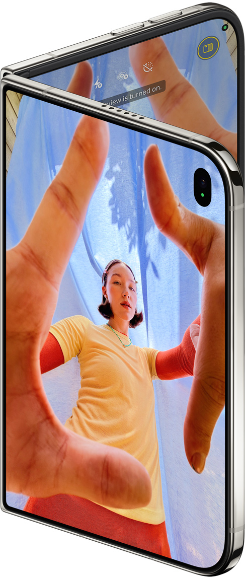 Lekko otwarty iPhone Duo w kolorze gwiezdnej bieli, na wyświetlaczu zewnętrznym widać aplikację Aparat i aktywny przedni aparat Center Stage, a na wyświetlaczu wewnętrznym bibliotekę zdjęć w aplikacji Zdjęcia