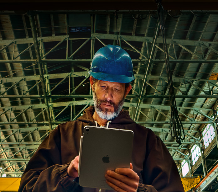 iPad i silver med en Apple Pencil som sitter fast, hållen av en person i en brun jacka och en blå hatt, som arbetar i en industriell miljö med metallbalkar ovanför