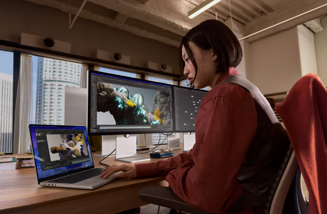 En person sitter vid ett skrivbord med en MacBook Pro ansluten till en extern skärm, båda skärmarna visar en 3D-rendering, byggnader syns genom fönstret bakom