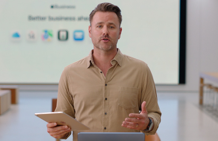 Presentatören talar och hänvisar samtidigt till en iPad och en stor skärm visar ikoner för olika företagsappar.