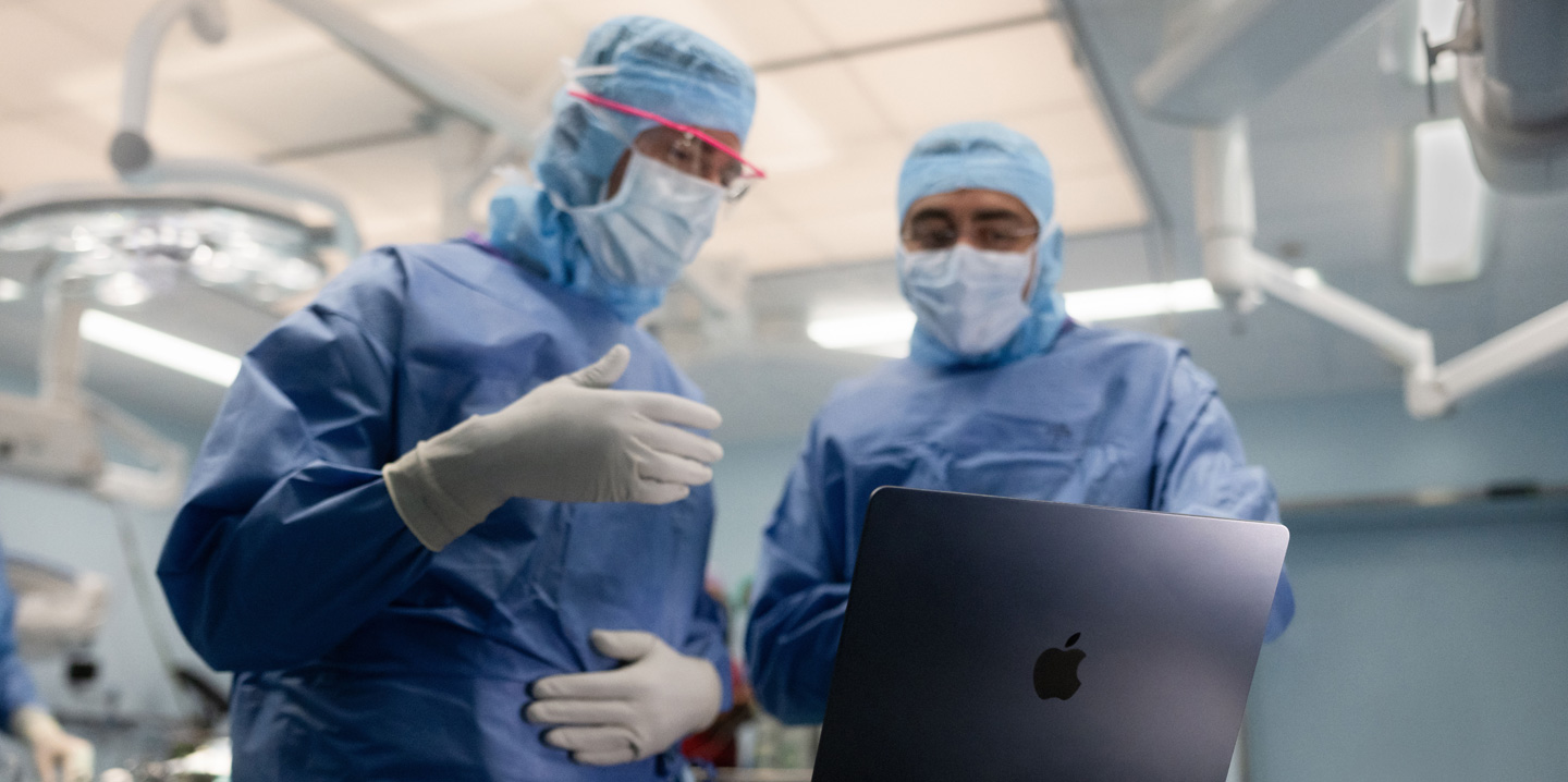 Hai bác sĩ phẫu thuật trong phòng mổ nhìn vào MacBook, một người ra hiệu bằng tay trong khi người kia quan sát