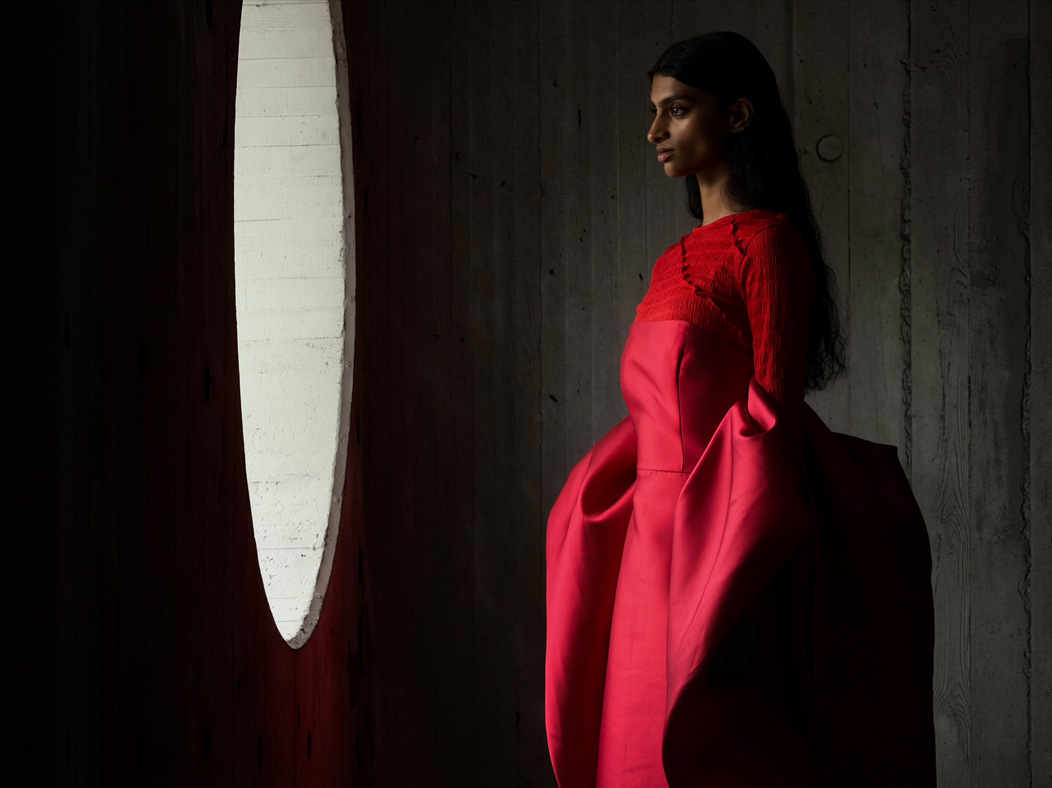 Uma mulher de vestido vermelho longo está sendo fotografada em um ambiente vazio. O app Fotos alterna entre as distâncias focais para demonstrar os recursos do sistema de câmera Pro, mostrando: 200 mm