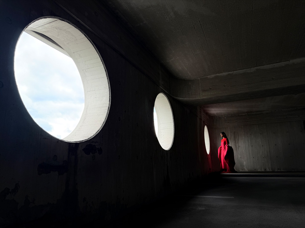 Uma mulher de vestido vermelho longo está sendo fotografada em um ambiente vazio. O app Fotos alterna entre as distâncias focais para demonstrar os recursos do sistema de câmera Pro, mostrando: 24 mm