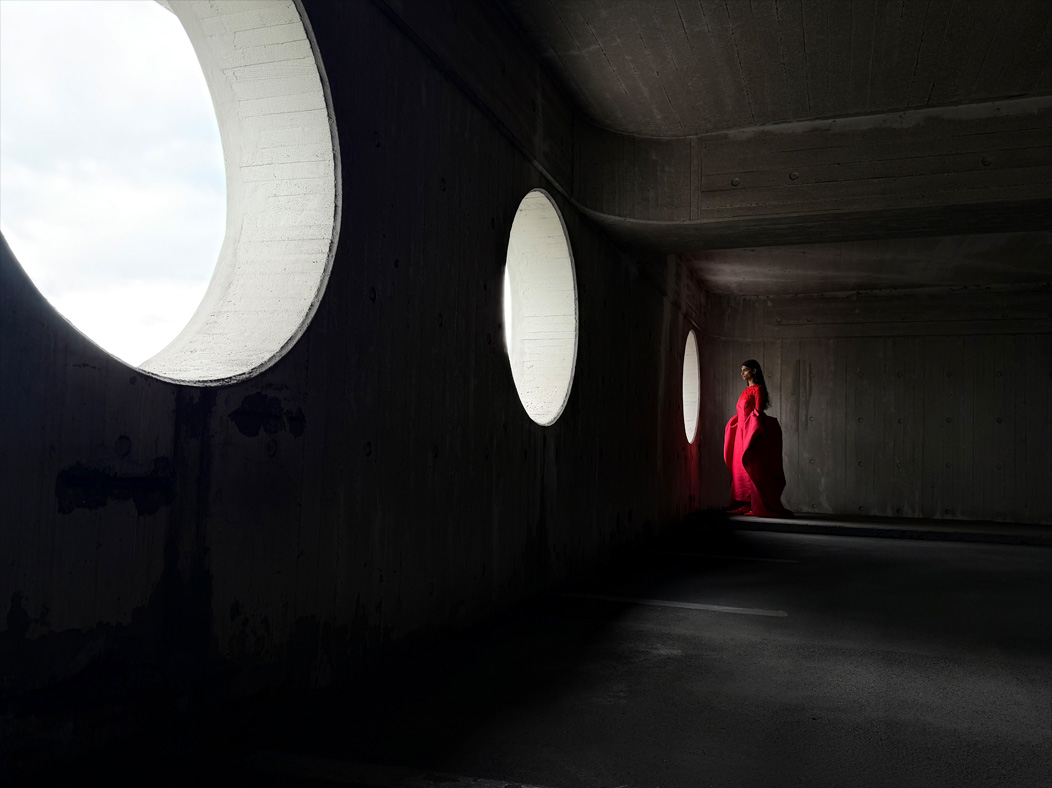 Uma mulher de vestido vermelho longo está sendo fotografada em um ambiente vazio. O app Fotos alterna entre as distâncias focais para demonstrar os recursos do sistema de câmera Pro, mostrando: 28 mm
