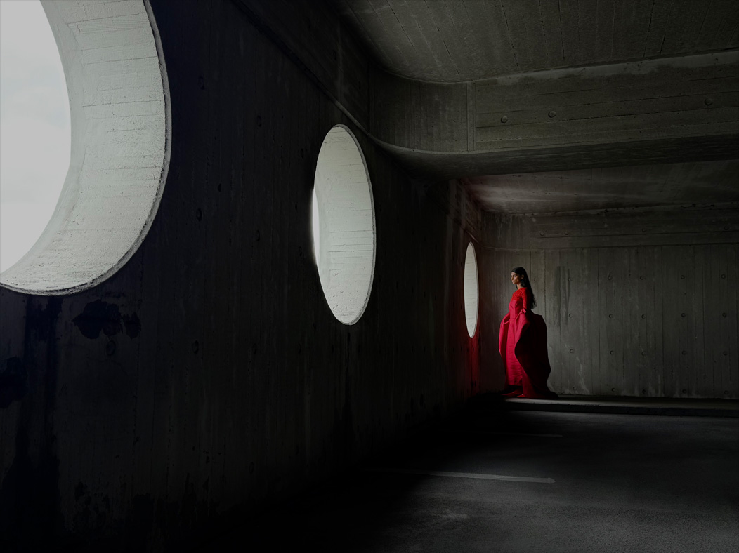 Uma mulher de vestido vermelho longo está sendo fotografada em um ambiente vazio. O app Fotos alterna entre as distâncias focais para demonstrar os recursos do sistema de câmera Pro, mostrando: 35 mm