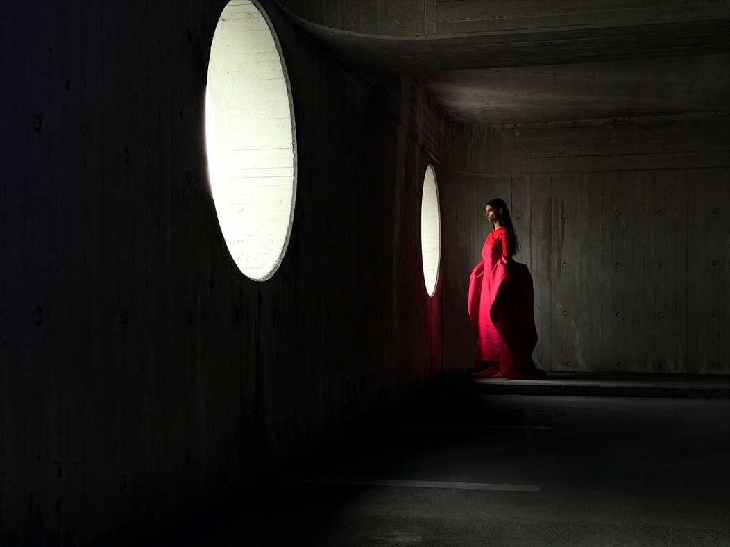Uma mulher de vestido vermelho longo está sendo fotografada em um ambiente vazio. O app Fotos alterna entre as distâncias focais para demonstrar os recursos do sistema de câmera Pro, mostrando: 48 mm