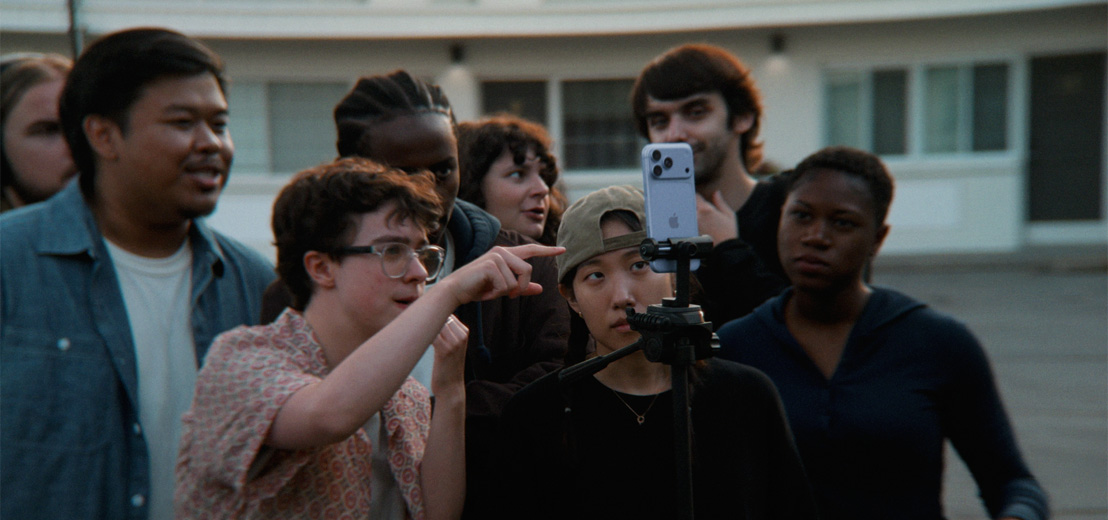 Two young filmmakers direct a scene to group of young people using an iPhone 18 Pro in Glacier colour mounted on a tripod