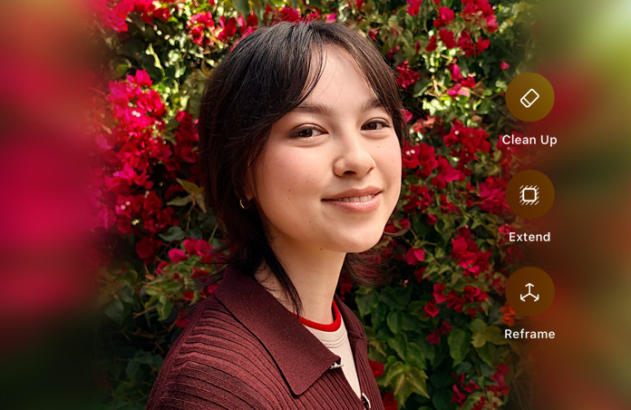 Photo of smiling person with brown hair, wearing a brown sweater, surrounded by red flowers, AI photo editing demo with Clean Up, Extend, Reframe options shown over photo