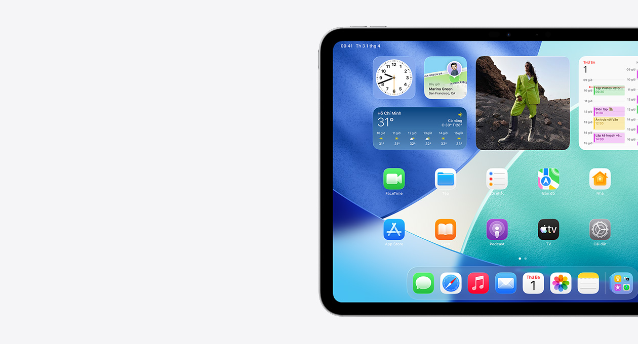 iPad nằm ngang, chỉ thấy phía bên trái màn hình, Màn Hình Chính với các tiện ích xếp chồng lên nhau bao gồm đồng hồ trong mờ thể hiện hiệu ứng Liquid Glass, ứng dụng Tìm, tiện ích Thời Tiết, hai hàng ứng dụng nhỏ hơn và dock